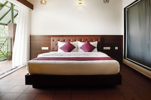 Una cama bien hecha con tablero de madera, almohadas blancas y moradas, y una manta roja.