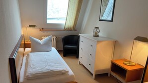 Et soveværelse med seng, natbord, kommode, stol og et vindue med gardiner.