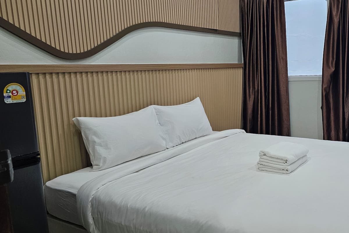 A neatly made bed with white linens and a folded towel, a wooden headboard, and a view of a green fence through a window with brown curtains.