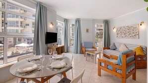 A modern living room with a dining area, large windows, a flat-screen TV, and a sofa with cushions. - Colmeia AL - Casa Abelha: Apartamento a poucos passos da praia em Armação de Pera (Armação de Pêra)