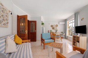 Un salon comprenant un canapé, des fauteuils, une table basse, un téléviseur, un coin repas avec des chaises, une porte en bois et une fenêtre avec des rideaux.