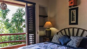 Un dormitorio con una cama, una mesita de noche, una lámpara y vistas al verdor por una puerta abierta.