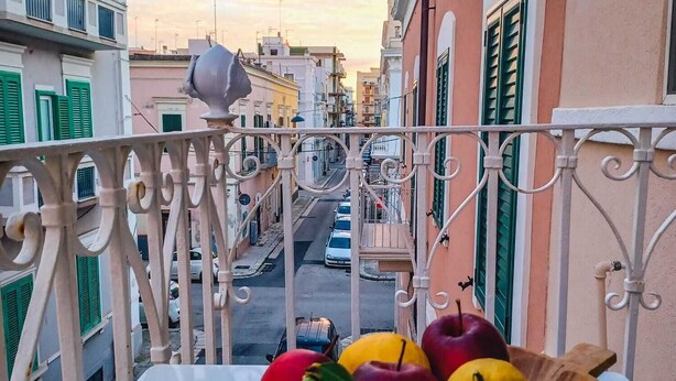 A balcony with a view of a street, a table with fruits, and buildings with green shutters. - GIONI House (Monopoli)