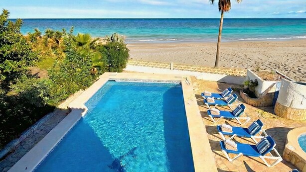 A swimming pool with clear blue water, surrounded by lounge chairs and a palm tree, overlooking a beach and ocean. - Villa on the beach with private pool - holaVivienda Inmobiliaria (Dénia)