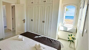 A bedroom with a bed, a wardrobe, a window with a view of the beach, and a vase with flowers. - Villa on the beach with private pool - holaVivienda Inmobiliaria (Dénia)