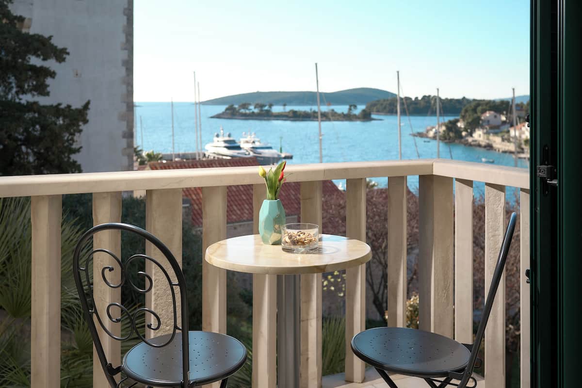 Een balkon met twee stoelen, een kleine tafel met een vaas en een schaal met noten, en uitzicht op zee met boten en een eiland in de verte.