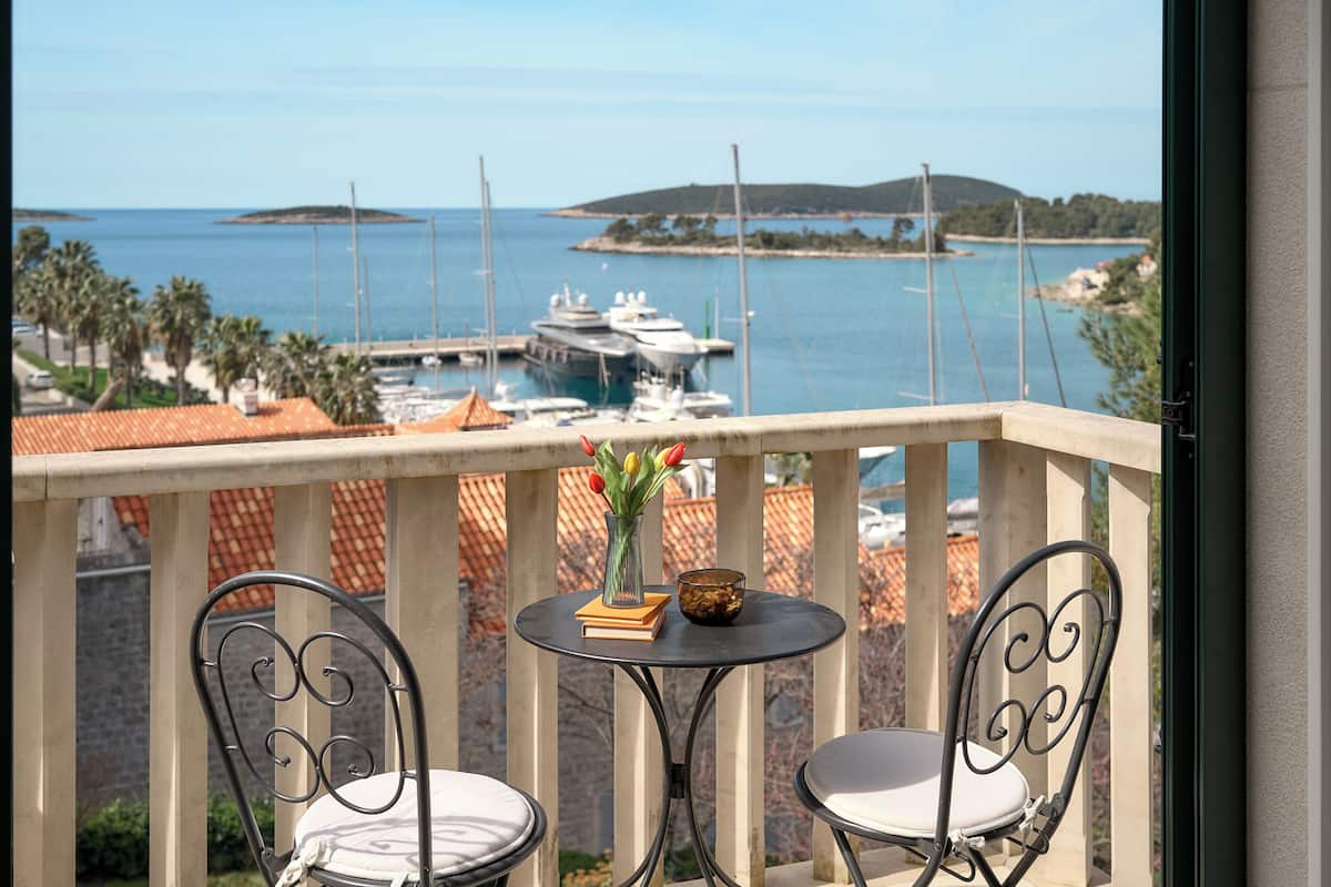 Een balkon met twee metalen stoelen en een kleine ronde tafel, met uitzicht op een jachthaven vol boten en een eiland in de verte.