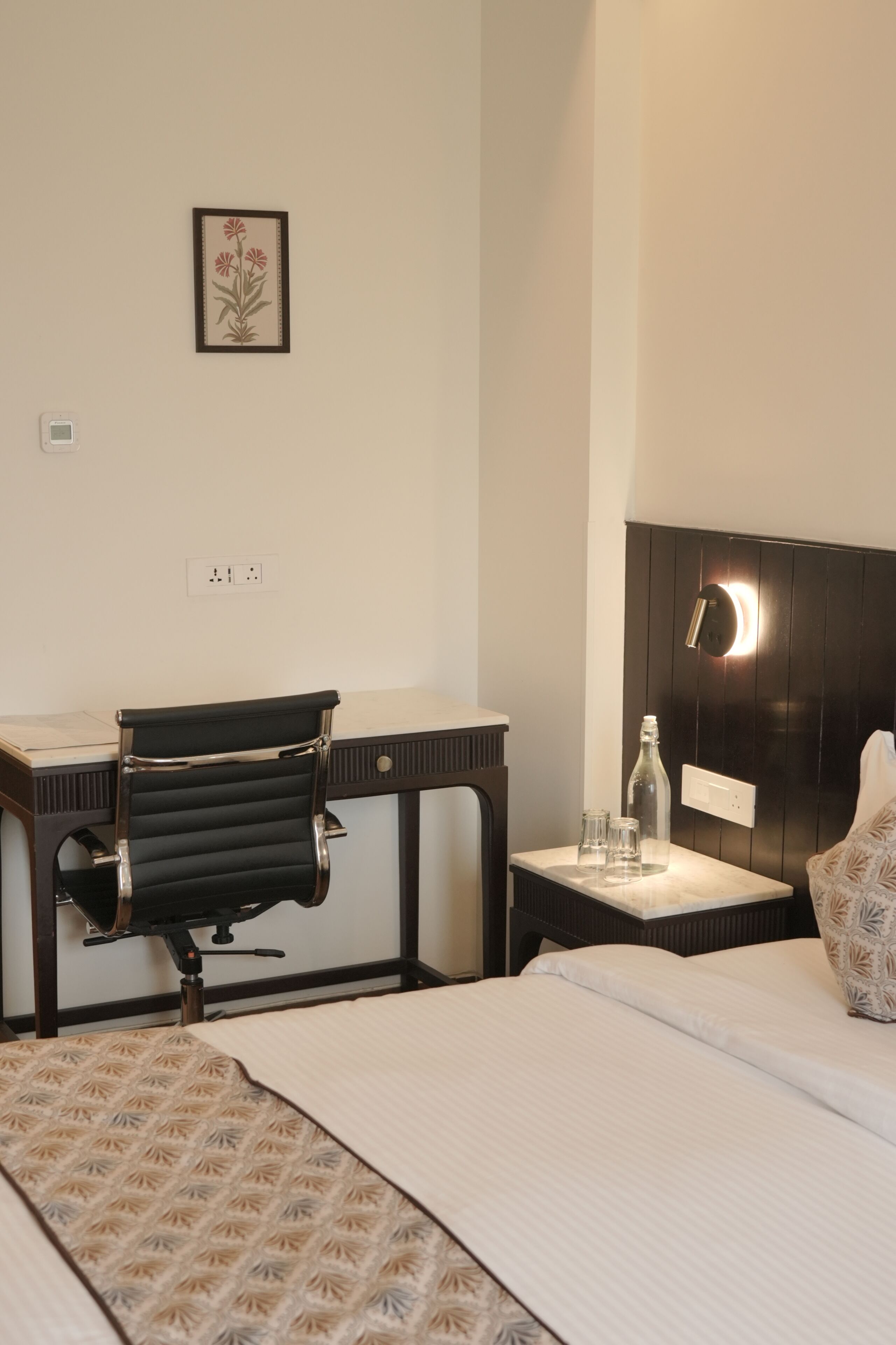 A hotel room with a bed, a desk with a chair, a bedside table with a lamp, and a wall-mounted outlet.