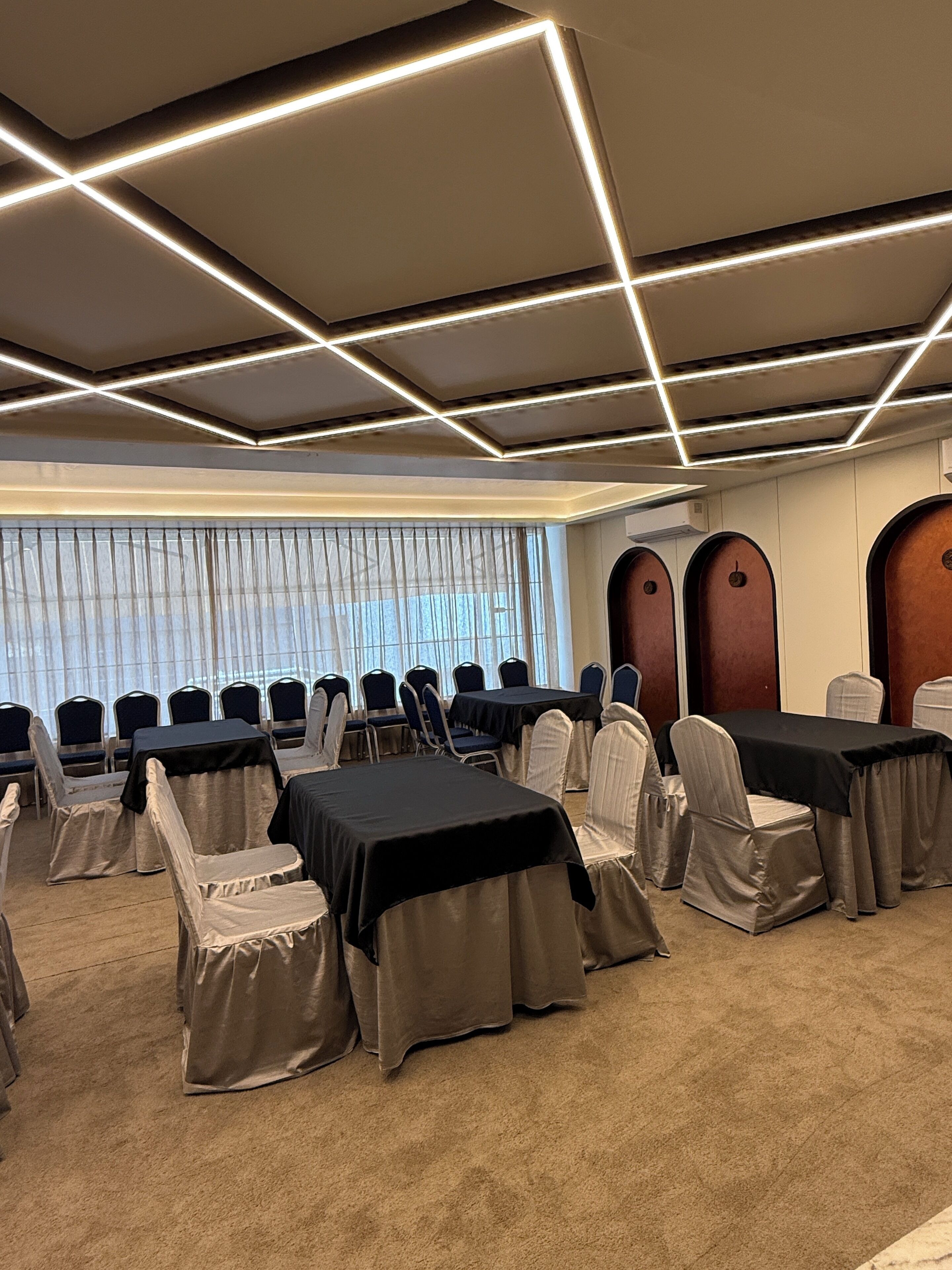 A conference room with tables and chairs, a ceiling with linear lighting, and large windows with blinds.