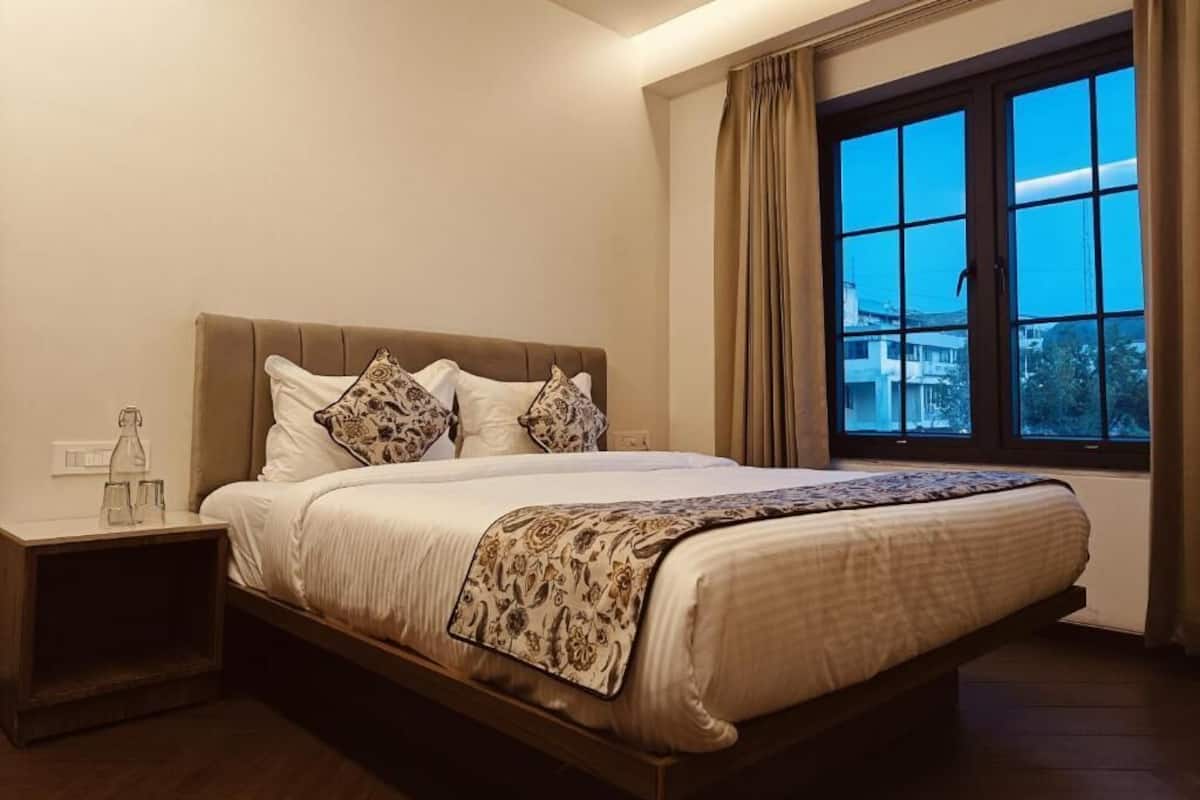 A neatly made bed with a patterned bedspread, a bedside table with a bottle and glasses, and a window with curtains.