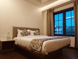 A neatly made bed with a patterned bedspread, a bedside table with a bottle and glasses, and a window with curtains. - Divik Aparthotel (Bhopal)