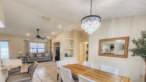 A modern dining area with a wooden table and chairs, a chandelier, and a view into the living room. - Solar Heated Pool, Sleeps 10, Peaceful retreat close to shopping and beach! (Palm Coast)