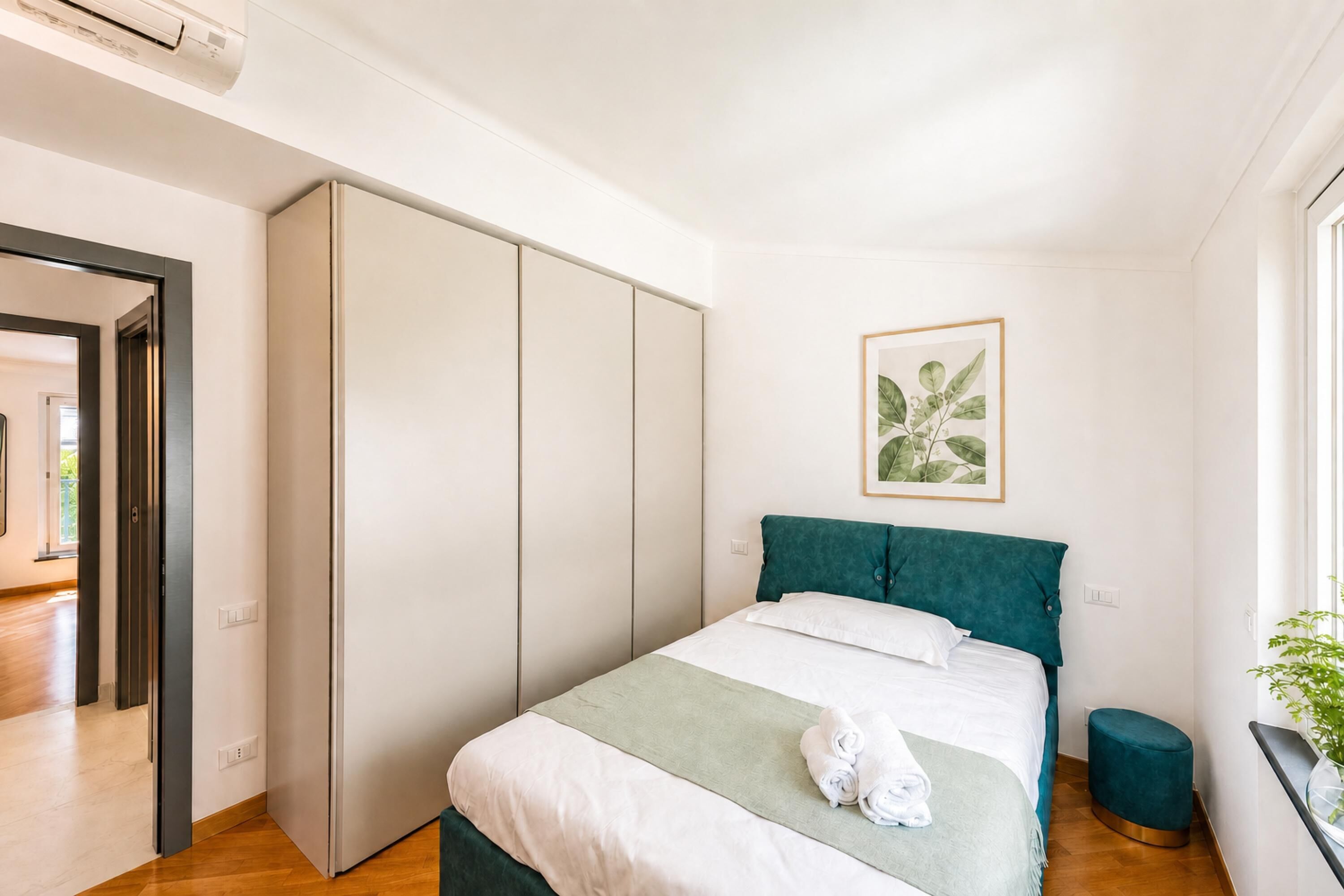 A bedroom with a bed, a wardrobe, a bedside table, a window, and a plant.