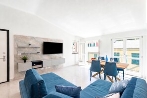 Un salon moderne avec un téléviseur à écran plat fixé sur un mur de marbre, un coin repas avec une table en bois et des chaises bleues, et un balcon avec vue.