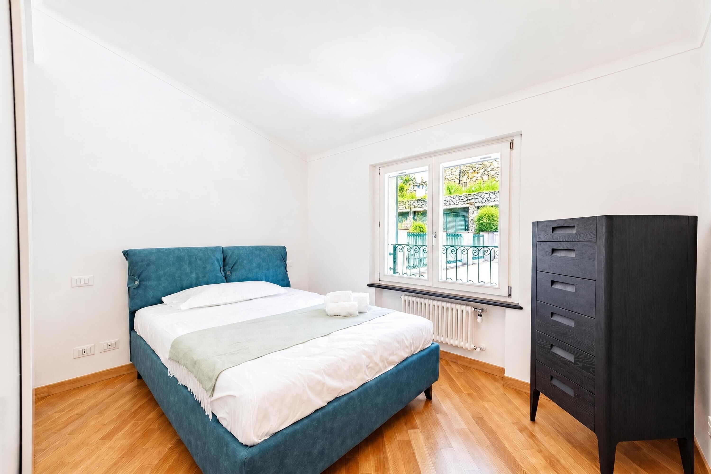 A bedroom with a bed, a radiator, a window with a view, and a black wardrobe.