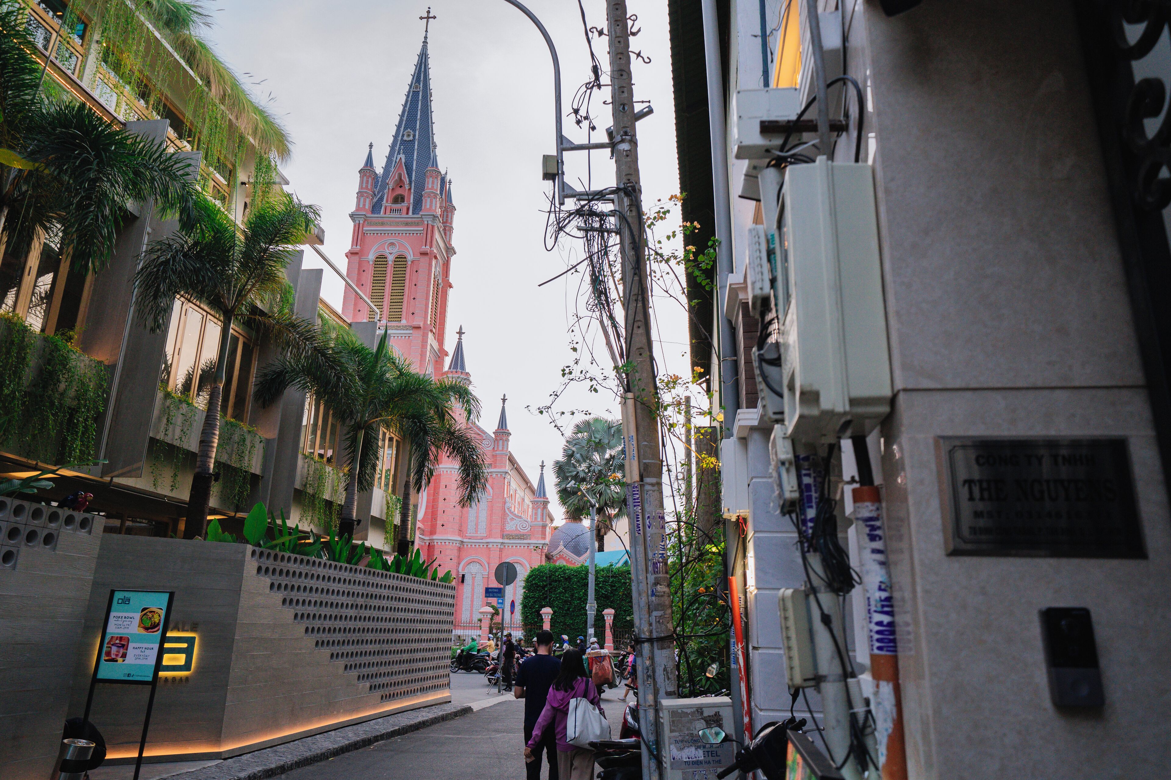 Vista callejera con una iglesia rosa al fondo, un edificio moderno con vegetación a la izquierda y un cartel que dice "The Nguyen" a la derecha.
