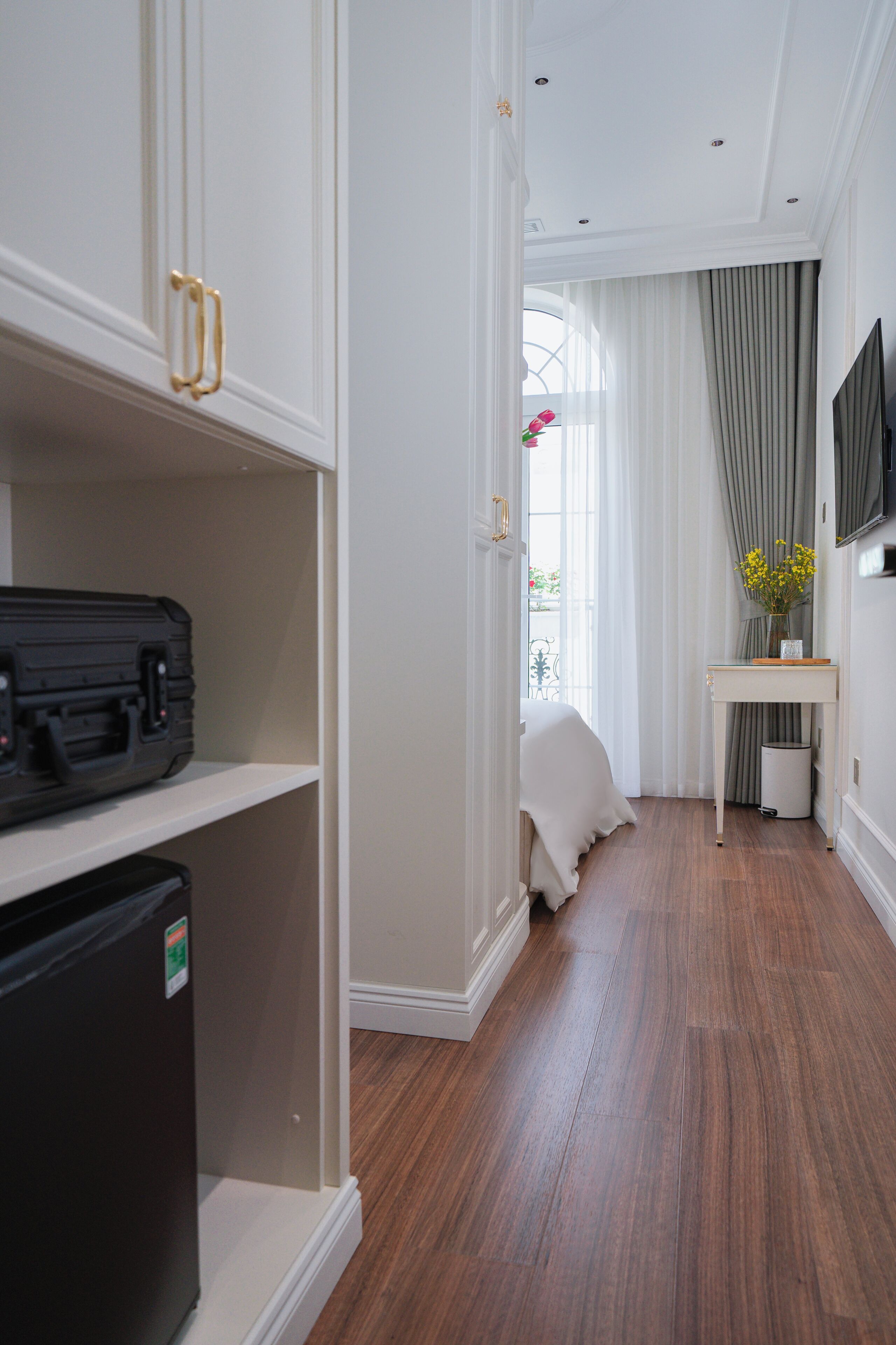 A hotel room with a wooden floor, a bed with white bedding, a desk with a vase of flowers, and a black suitcase on a shelf.