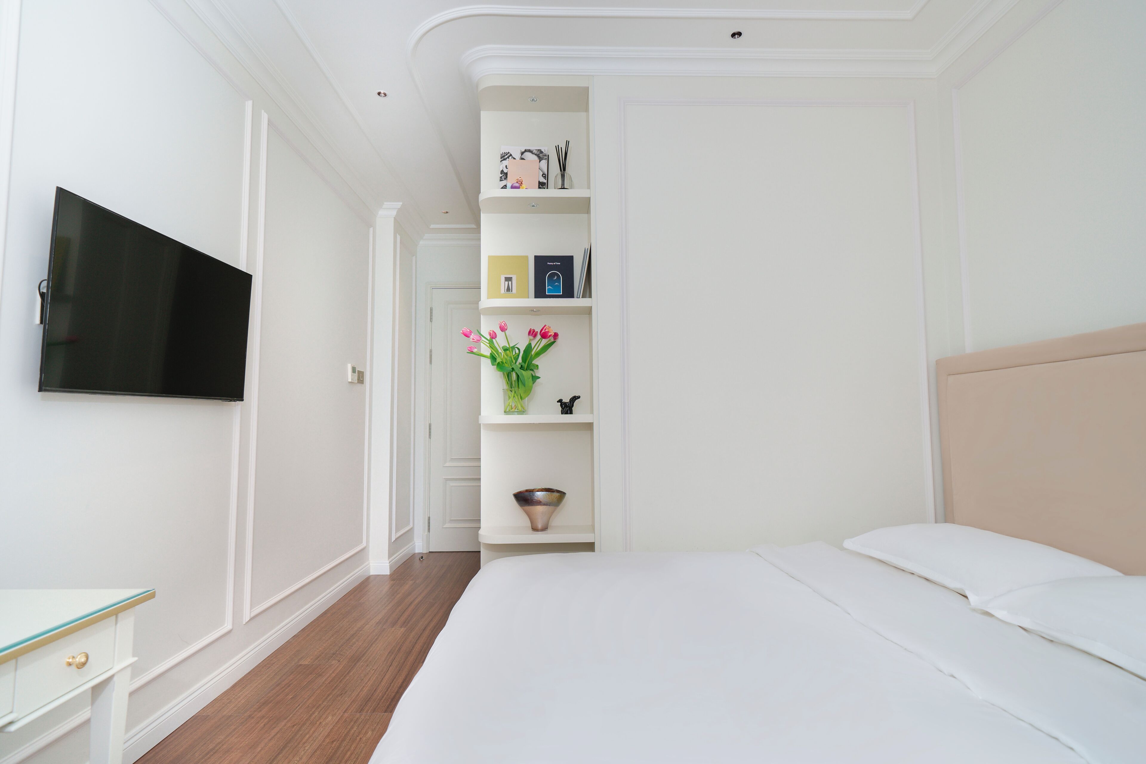 A bedroom with a bed, a TV mounted on the wall, a bedside table, and a built-in shelving unit with decorative items.
