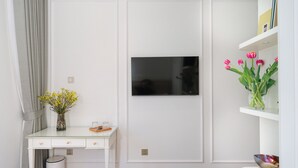 Une chambre à coucher avec un lit, un bureau agrémenté d’un vase de fleurs, un téléviseur fixé au mur et une étagère avec des livres et un vase de tulipes.