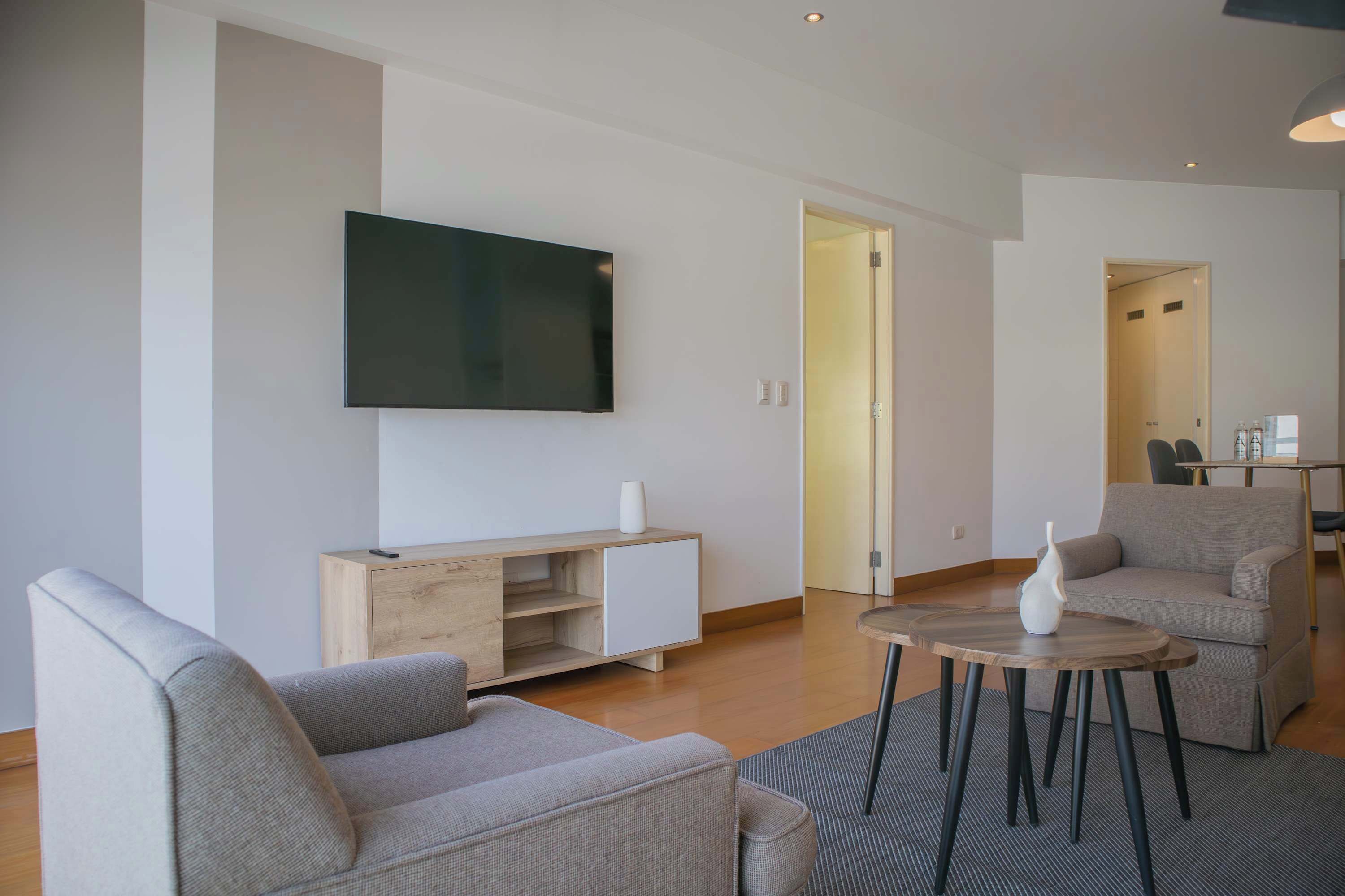 A modern living room with a flat-screen TV mounted on the wall, a wooden cabinet, a grey sofa, a round coffee table, and a grey armchair.