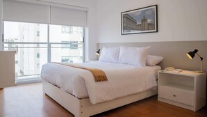 Quarto de hotel moderno com uma cama grande, uma mesinha de cabeceira com abajur, um quadro na parede e vista da cidade pela janela.