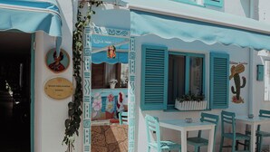 A building with a blue awning, a door with a circular sign, and a table with chairs outside. - Frida Sığacık Butik Otel (Seferihisar)