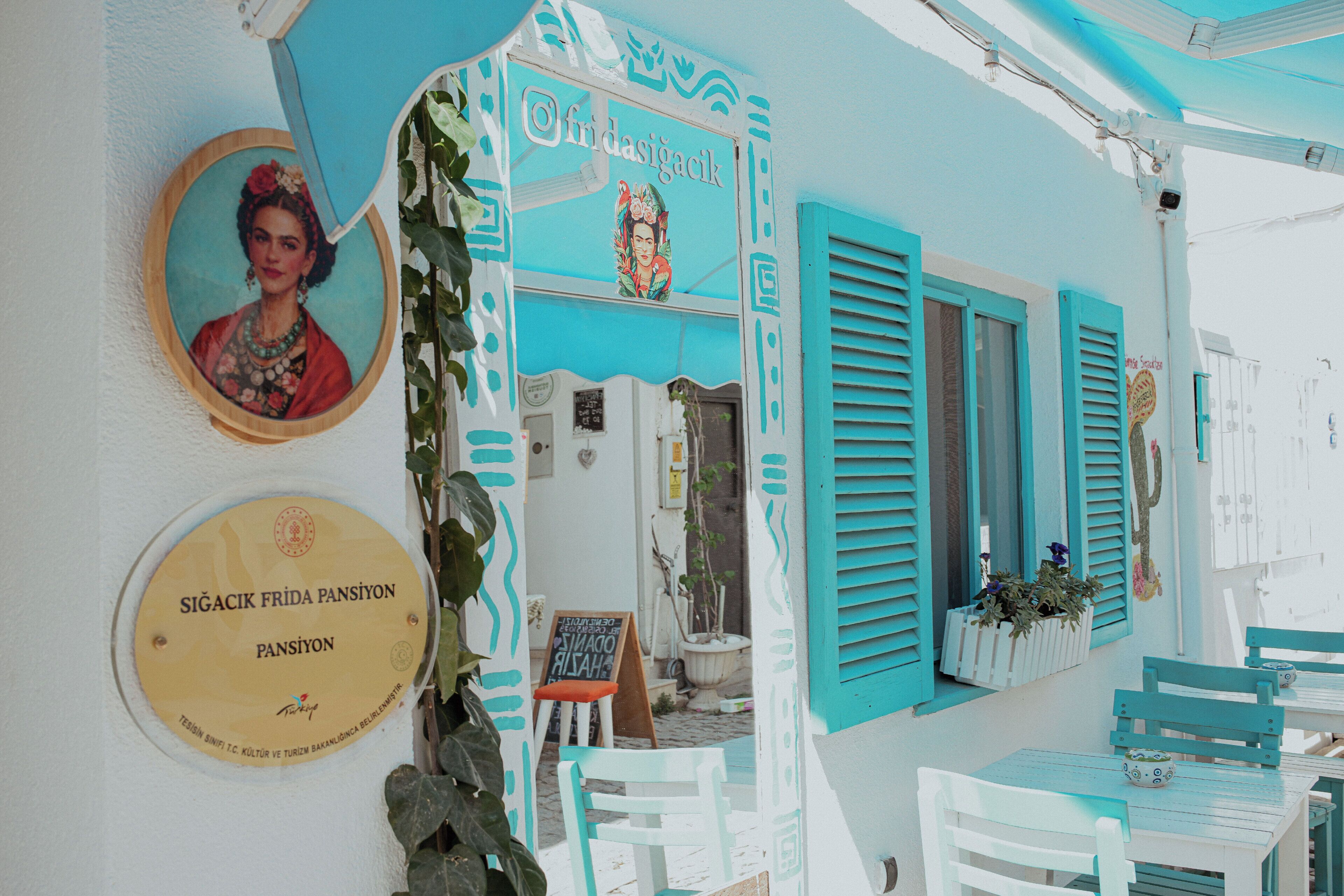 Un edificio con contraventanas turquesas y un letrero que dice 'Frida Pansiyon'. Hay una placa en la pared y un cuadro enmarcado.