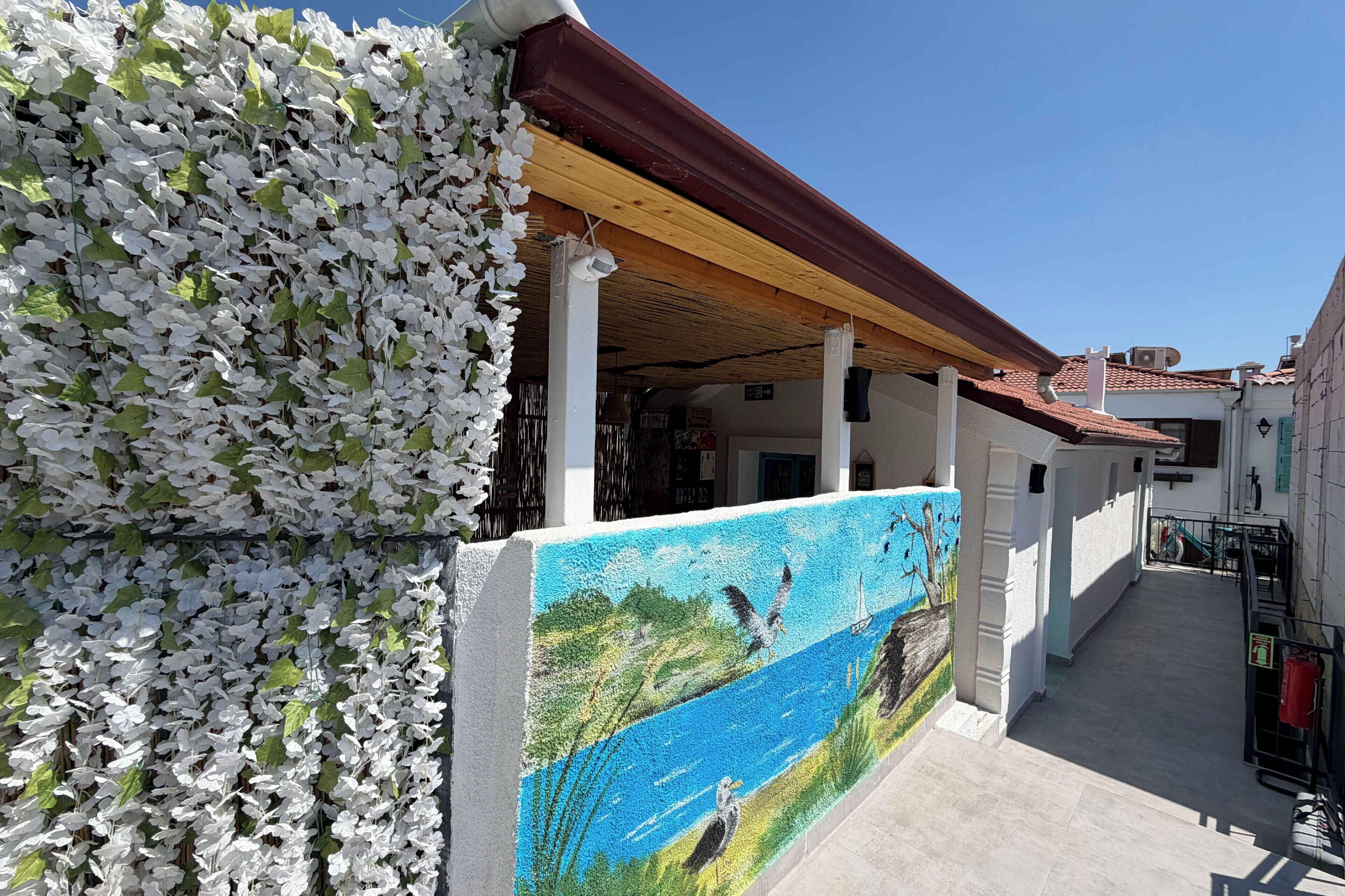 Un edificio con un paseo techado, un mural con aves y agua, y una pared cubierta de flores blancas.