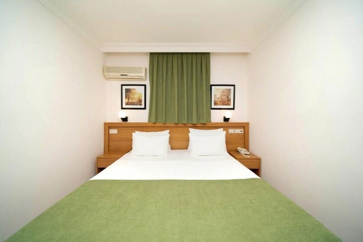 A hotel room with a single bed, a wooden headboard, a green bedspread, two bedside tables with lamps, a telephone, and two framed pictures on the wall.
