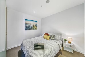 Un dormitorio con cama, almohadas, una mesita de noche con lámpara, una planta pequeña y un cuadro en la pared.