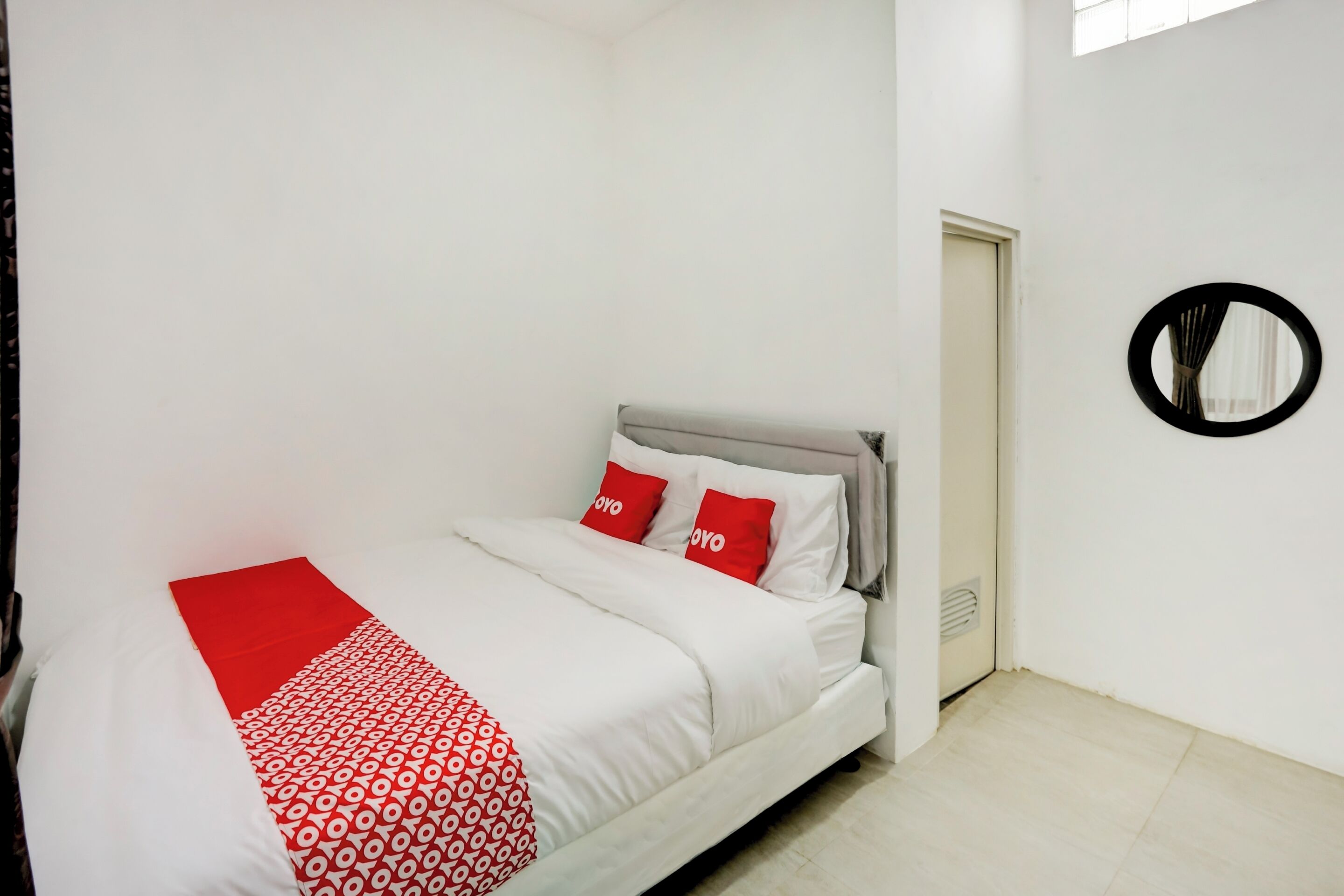 A neatly made bed with white and red bedding, a circular mirror on the wall, and a door to the right.