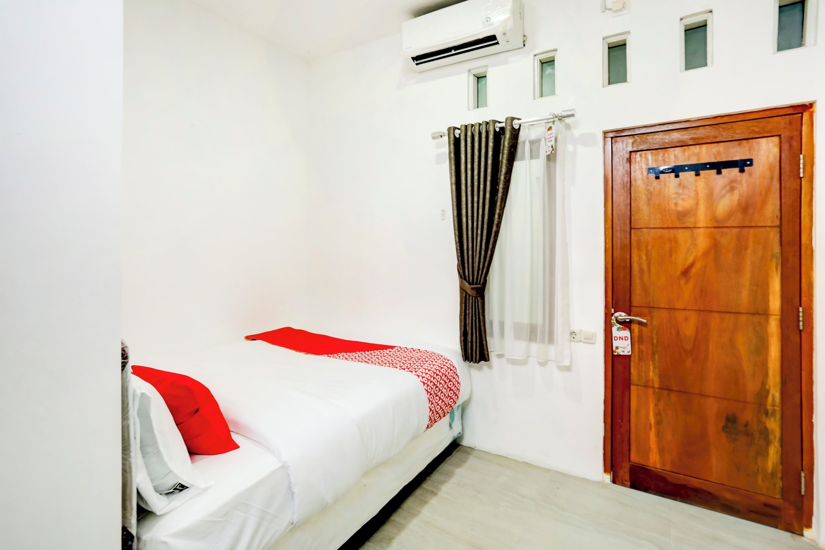 A small, clean room with a single bed, a wooden door, and an air conditioning unit on the wall.