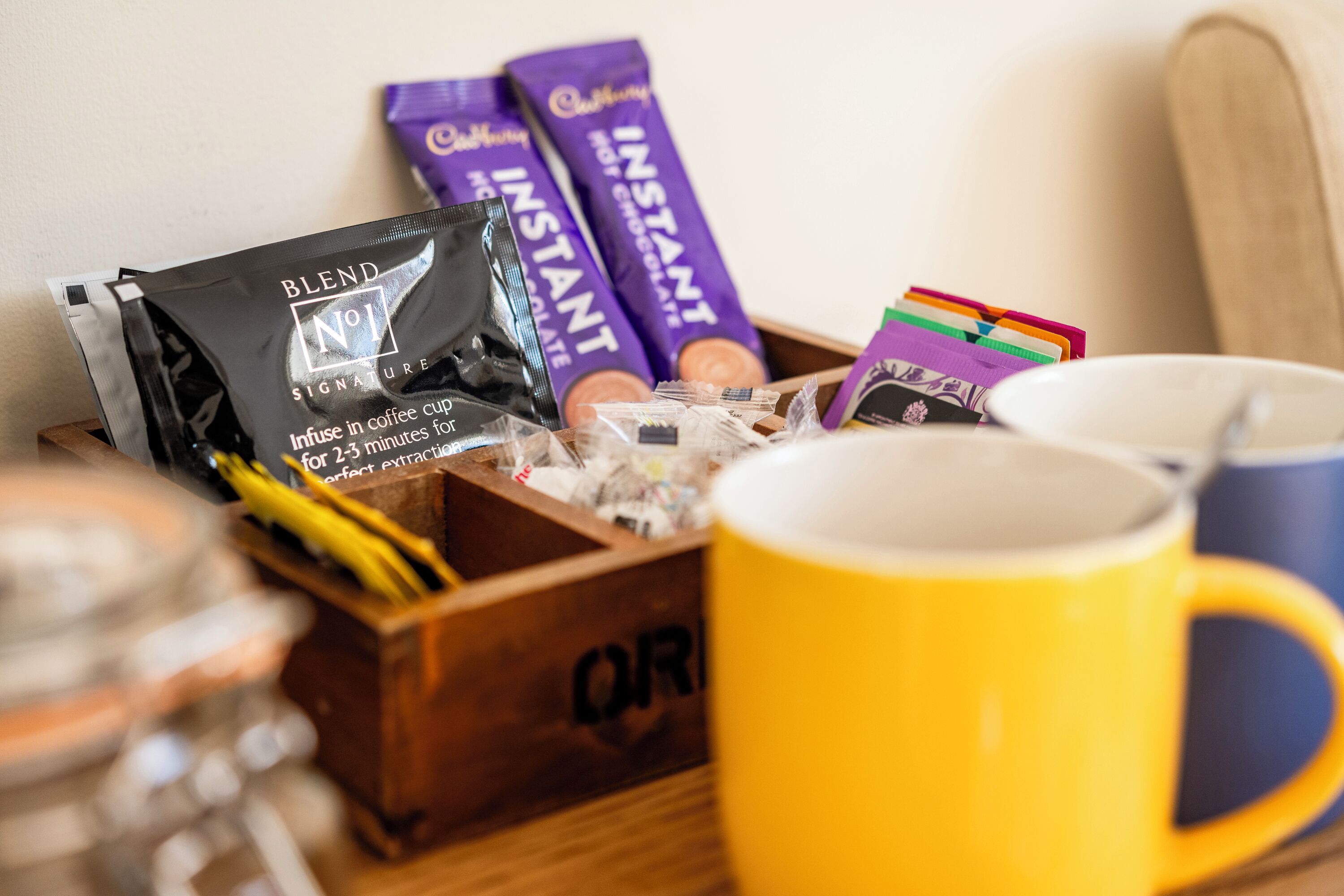 A wooden box with a yellow mug, two Cadbury chocolate bars, and a coffee pod.
