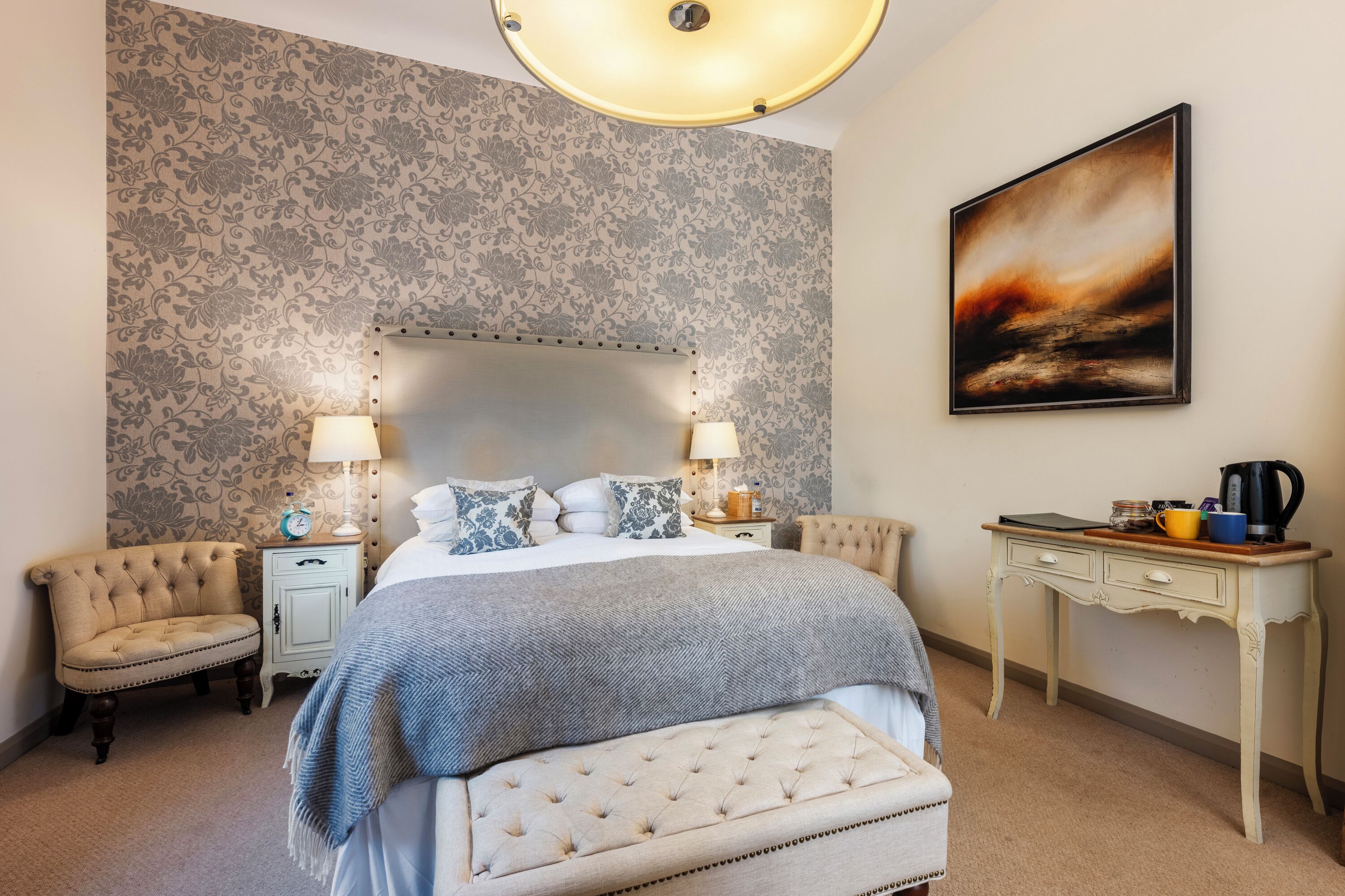 A bedroom with a large bed, two bedside tables, a nightstand, a bench, a desk, and a painting on the wall.