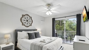 Ein modernes Schlafzimmer mit einem großen Bett, einem Deckenventilator, einem Fernseher und einem Balkon mit Blick auf Grünflächen.
