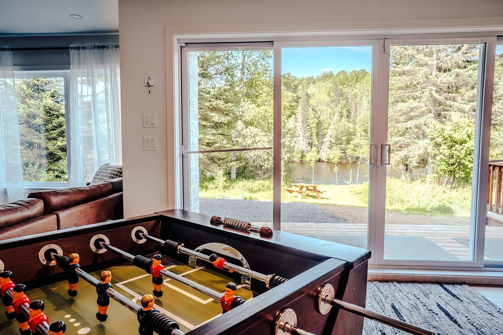 Una mesa de fútbolín en una habitación con un amplio ventanal corredero que da a una terraza con vistas a un lago y un bosque.