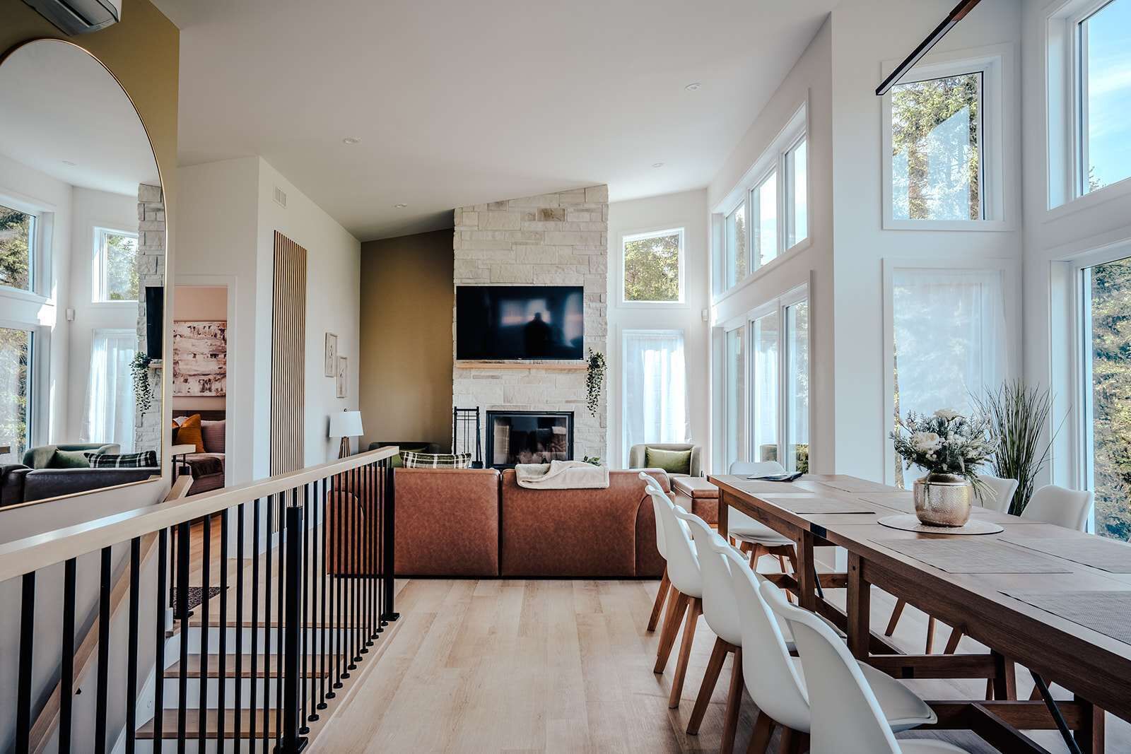 A modern living room with a fireplace, a dining area with a long table and chairs, and large windows providing natural light.