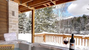 Un jacuzzi con una botella de vino y una copa en una terraza de madera con vistas a un paisaje nevado.