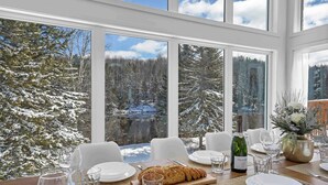Mesa servida con platos, vasos y una hogaza de pan, con vistas a un bosque nevado y un lago.