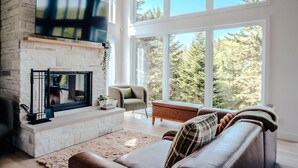 Una sala de estar con chimenea, televisor grande, sofá y vistas a árboles a través de amplios ventanales.