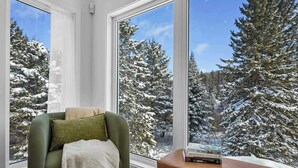 Una habitación acogedora con un sillón verde y una pequeña mesa con libros, con vistas a un bosque nevado a través de amplios ventanales.