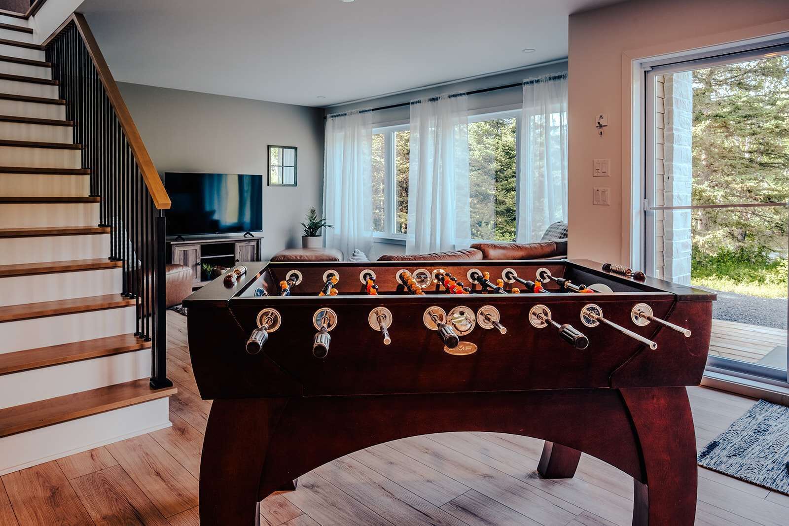 A modern living room with a foosball table, a staircase, a TV, and a large window with curtains.