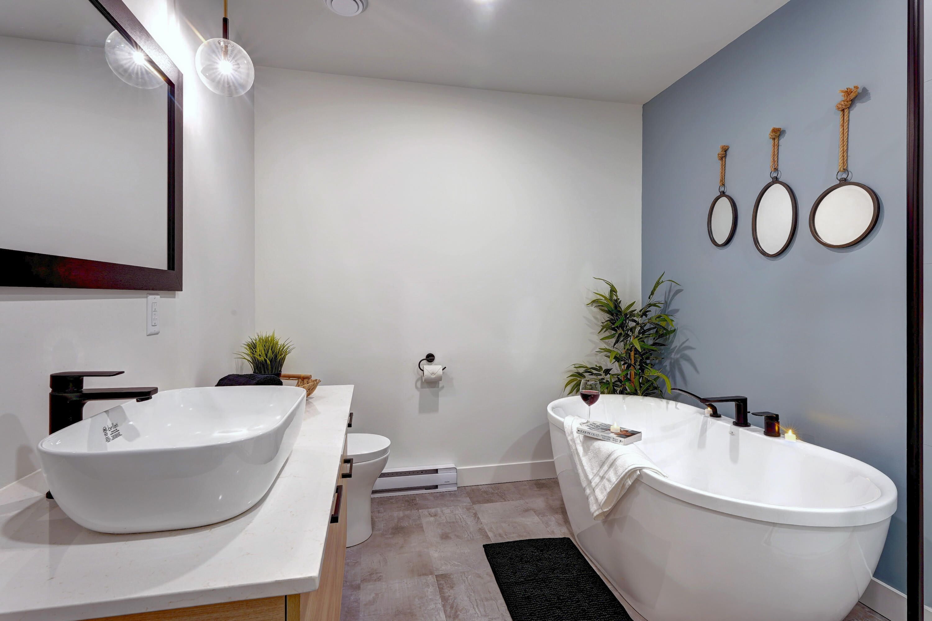 A modern bathroom with a white sink, a freestanding bathtub, and a toilet. There are three wall-mounted mirrors and a potted plant.