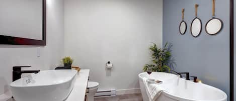 A modern bathroom with a white sink, a freestanding bathtub, and a toilet. There are three wall-mounted mirrors and a potted plant.