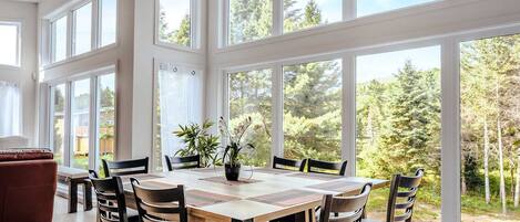 Uma sala de jantar com uma mesa grande, cadeiras e vista para uma floresta através de janelas amplas.