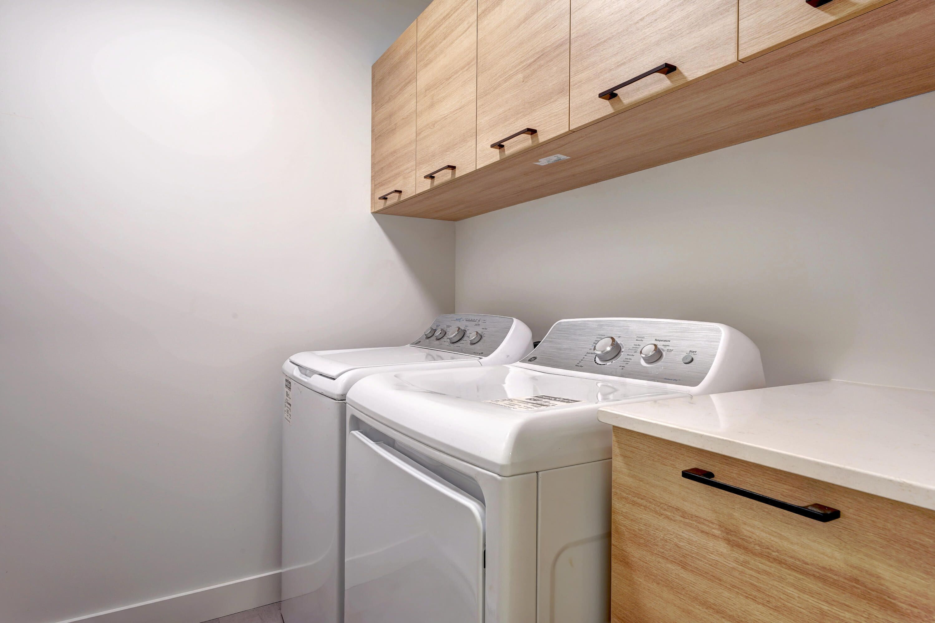 Un espace buanderie comprenant deux machines à laver blanches, un meuble en bois au-dessus et un plan de travail avec un tiroir.