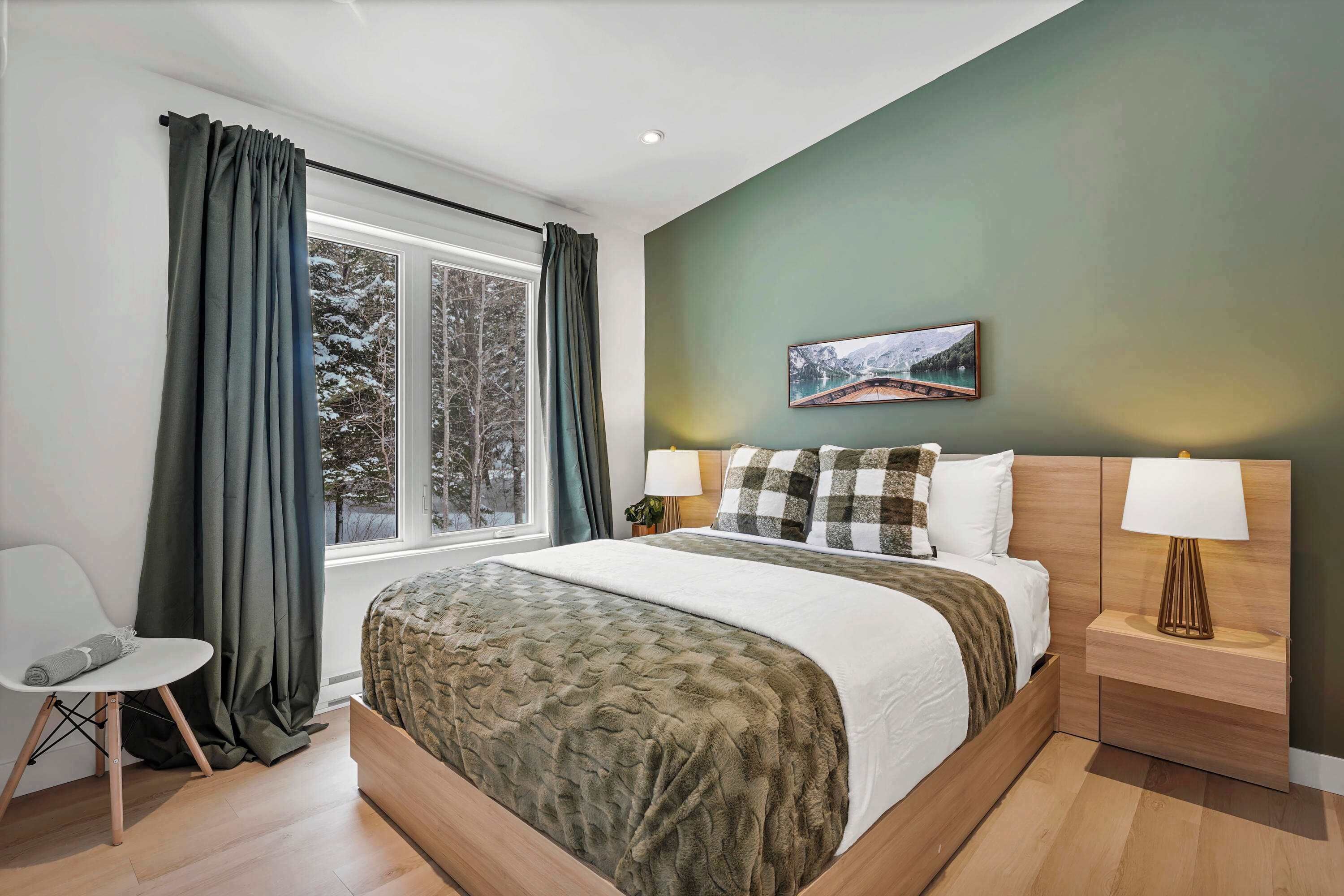 A bedroom with a large bed, a nightstand with a lamp, a window with curtains, and a painting on the wall.