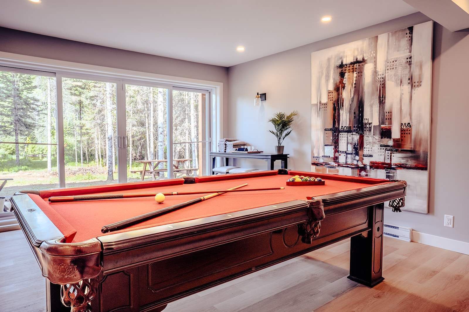 A pool table with balls, a potted plant, and a cityscape painting on the wall.
