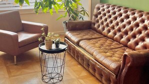 A living room with a leather sofa, a chair, a small table with a plant, and a view of a building through large windows. - hotel de la paix (Limoges)