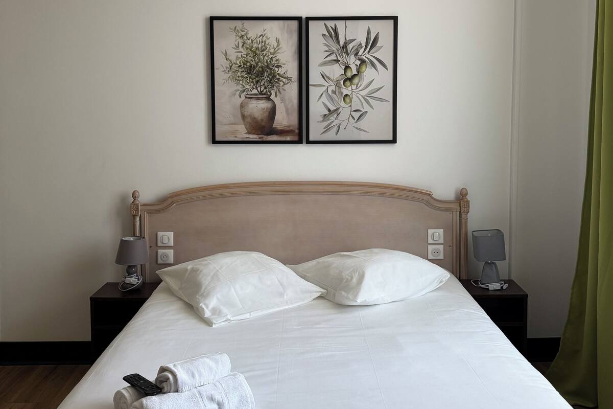 A neatly made bed with white linens, two white pillows, and a rolled white towel. There are two bedside tables with lamps and a wall-mounted light fixture. Two framed botanical prints are hung above the bed.